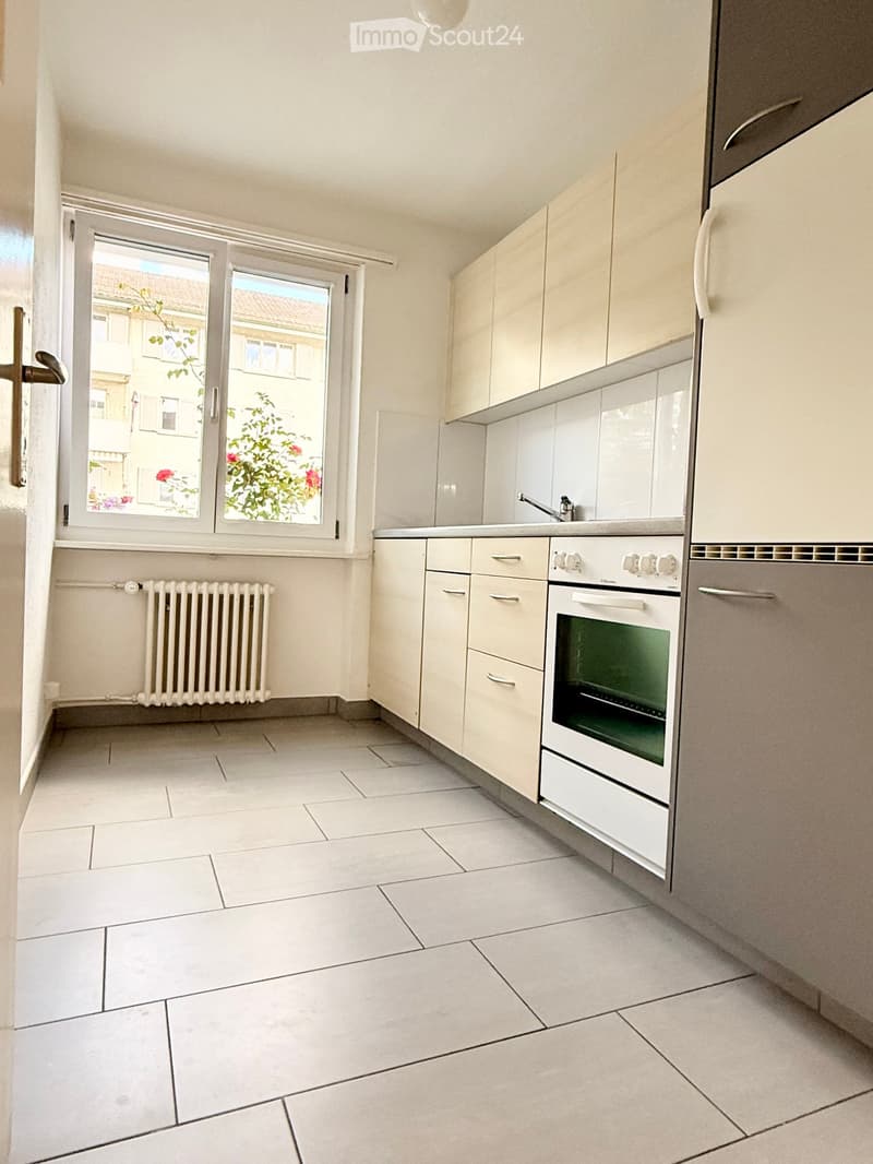 Modern kitchen with white cabinets, tiled floor, and a window overlooking a building with greenery.