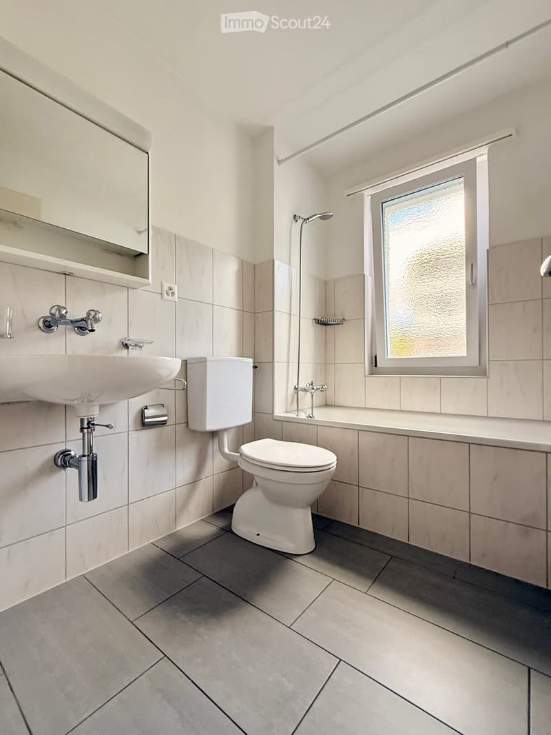 Modern bathroom with white tiles, a sink, toilet, bathtub, and a window with frosted glass.