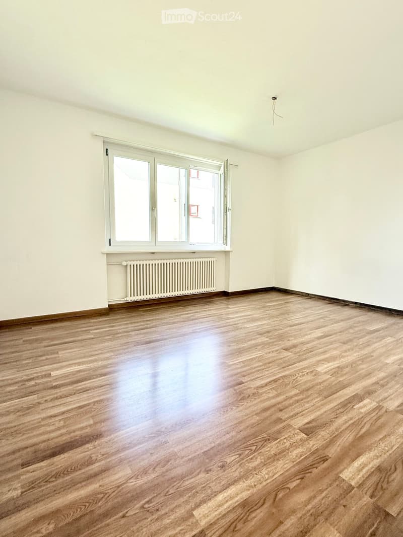 Empty room with wooden flooring, white walls, and a large window with a radiator below it.