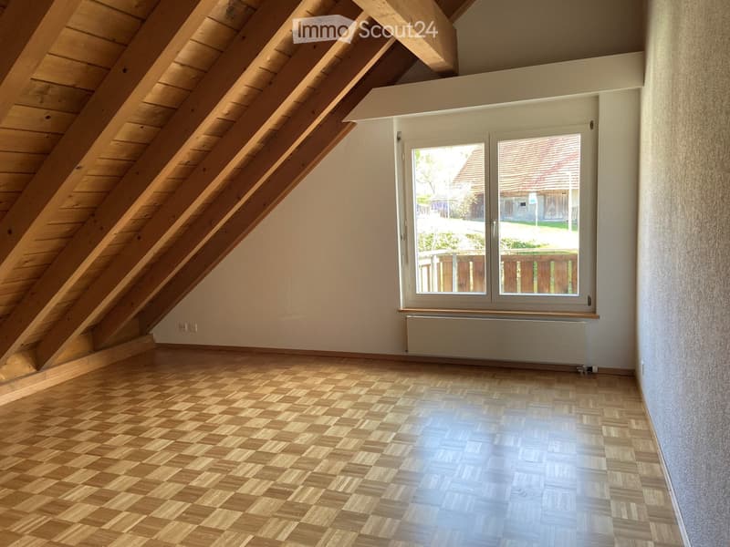 Empty attic room with wooden slanted ceilings, wooden flooring, and a large window offering a view of the outside. The room features a radiator below the window and exposed wooden beams.