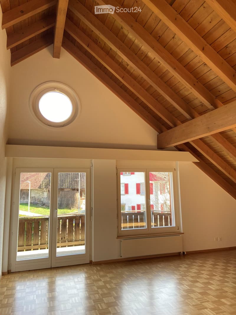Bright, empty attic room with wooden slanted ceilings, large windows, and a skylight. The floor is made of light wooden planks, and there is a radiator under one of the windows.