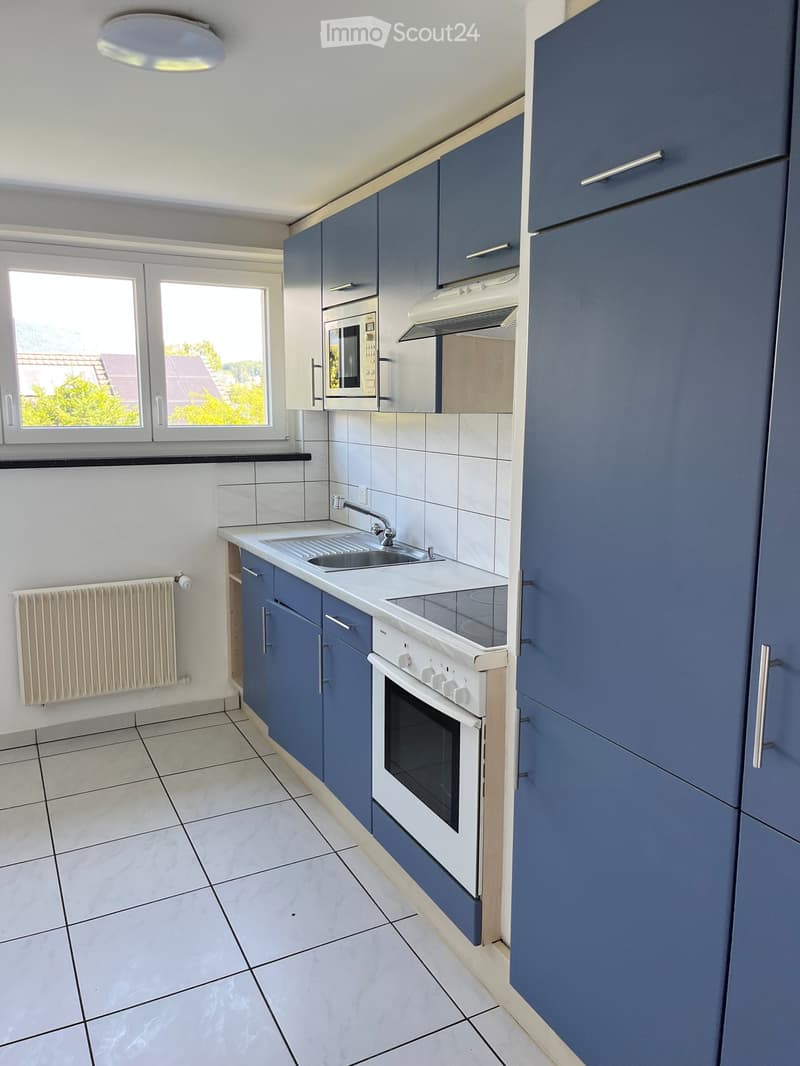 Modern kitchen with blue cabinets, white countertops, a sink, an oven, and a microwave. The space features tiled flooring and a window offering a view of greenery.