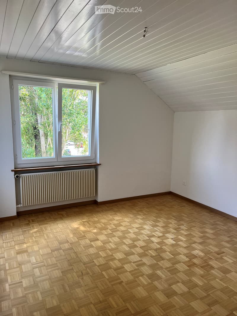 Empty room with wooden herringbone flooring, white walls, large window with view of greenery, radiator below window, and slanted white ceiling with visible beams.