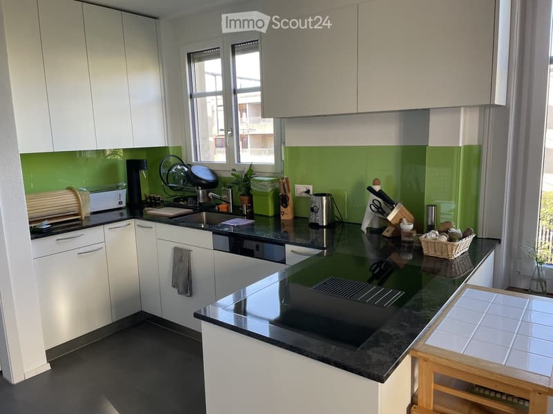 Modern kitchen with white cabinets, black countertops, and stainless steel appliances. Features a sink, coffee maker, toaster, blender, and a small plant on the counter.
