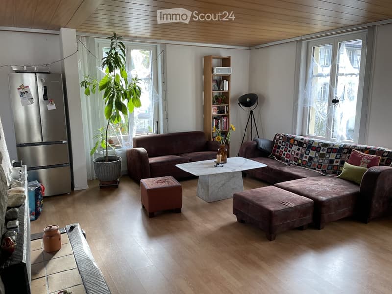 Bright living room with wooden flooring, large windows, a brown sectional sofa, a white coffee table, ottomans, a refrigerator, a bookshelf, a floor lamp, and a large potted plant.