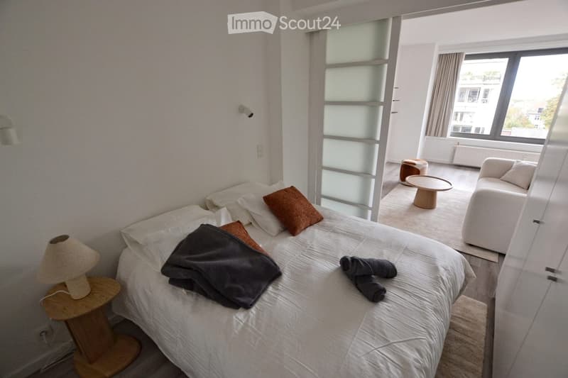 Modern bedroom with a neatly made bed, nightstand with lamp, a small sofa, a round wooden table, and a stool. Large windows with curtains provide natural light.