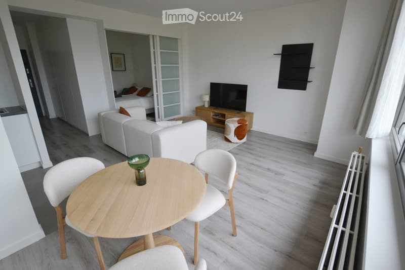 Modern living room with white sofa, small round wooden table with chairs, television on wooden stand, and a view into a bedroom with a bed and pillows.