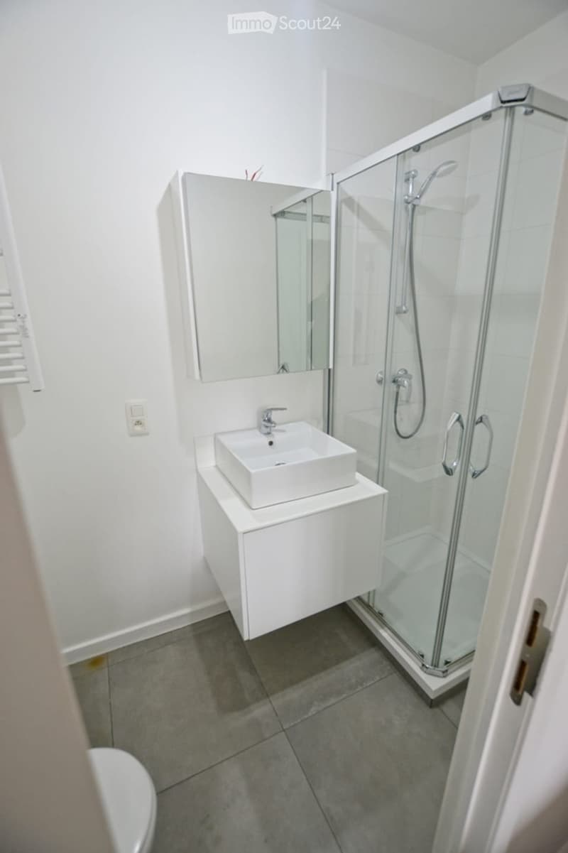 Modern bathroom with a white sink, mirror, and glass-enclosed shower. The floor is tiled, and the walls are white.