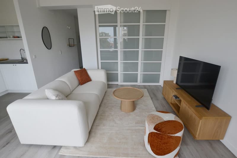 Modern living room with a white sectional sofa, wooden coffee table, round stool, flat-screen TV on a wooden stand, and a lamp. The room features large windows with frosted glass panels and a sleek, contemporary design.