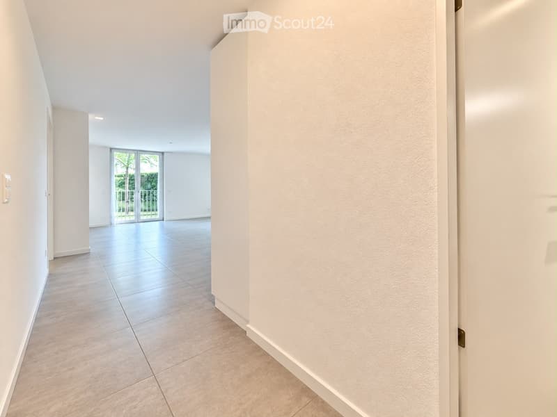 Empty hallway with tiled flooring, white walls, and a sliding glass door leading to a balcony area with outdoor views.