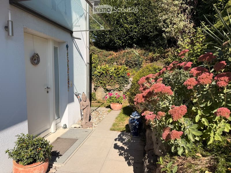A narrow garden path lined with potted plants and flowers, featuring a decorative statue and a small fountain, leading to a house entrance.