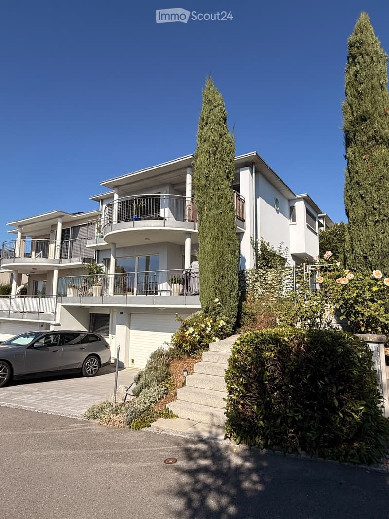 Modern two-story house with white exterior, multiple balconies, garage, and landscaped garden with tall coniferous trees and flowering shrubs.