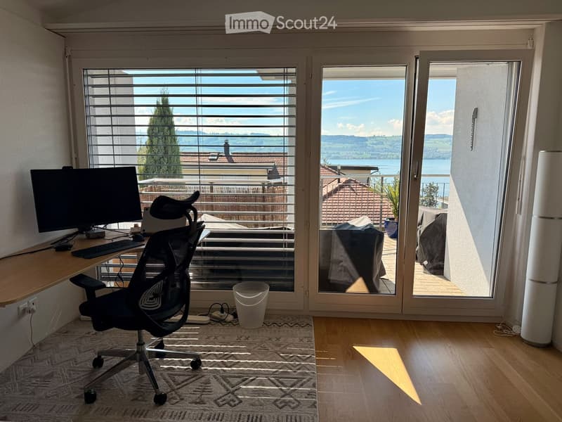 Bright room with large windows offering scenic views, equipped with a desk, computer, chair, and a trash bin.
