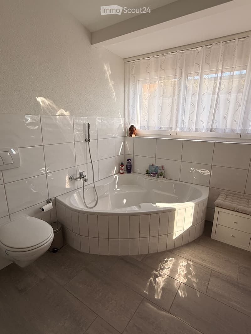 Modern bathroom with a large, corner bathtub, toilet, and small plants on the edge of the tub. The room features white tiled walls and a tiled floor, with a window covered by sheer curtains allowing natural light to enter.