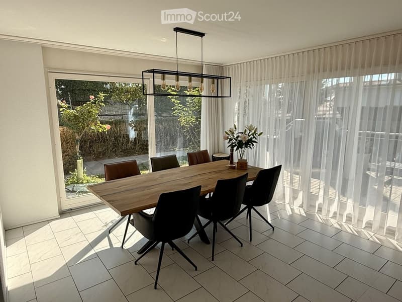 Modern dining room with a wooden dining table, six chairs, a pendant light fixture, and a vase with flowers on the table. Large windows with sheer curtains offer a view of the garden.