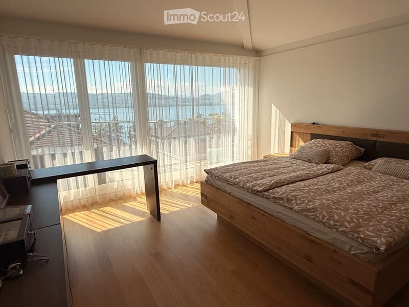 Bright bedroom with wooden flooring, large windows offering a scenic view, a double bed with patterned bedding, a nightstand, and a modern table with a chair.