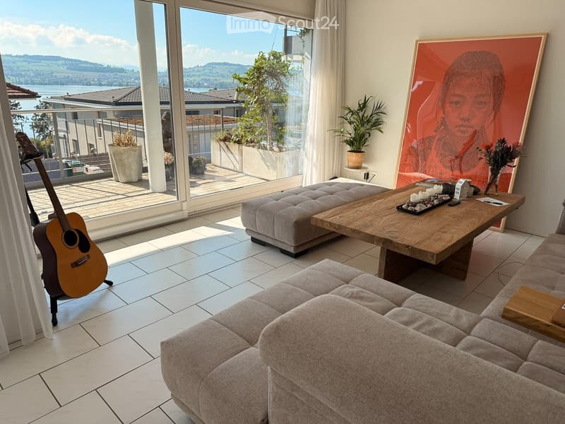 Modern living room with large windows offering a scenic view, featuring a grey sofa, wooden coffee table, guitar, and a large painting on the wall.
