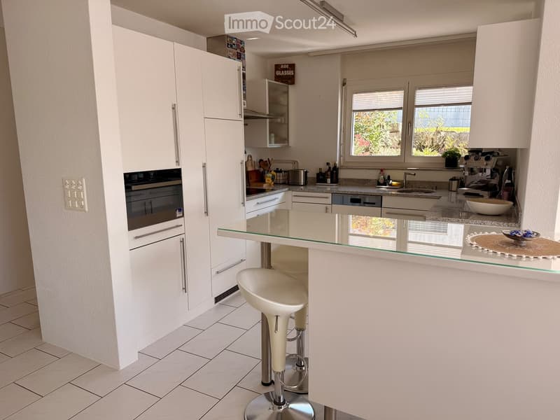Modern kitchen with white cabinets, stainless steel appliances, a glass island, and a stool. Features a window with a view of greenery.