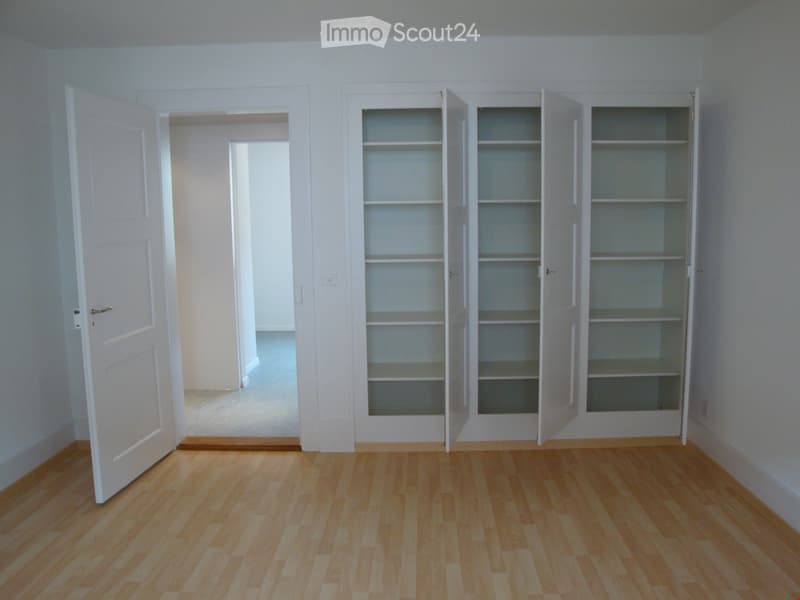 Empty room with light wooden flooring, white walls, and built-in white shelving units with glass doors. An open doorway leads to another room.