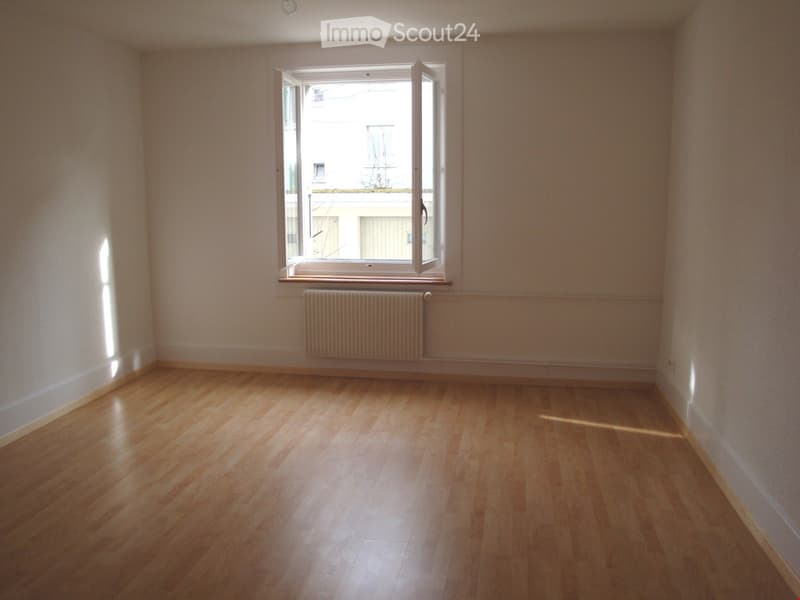 Empty room with wooden flooring, white walls, and a large window with a radiator below it.