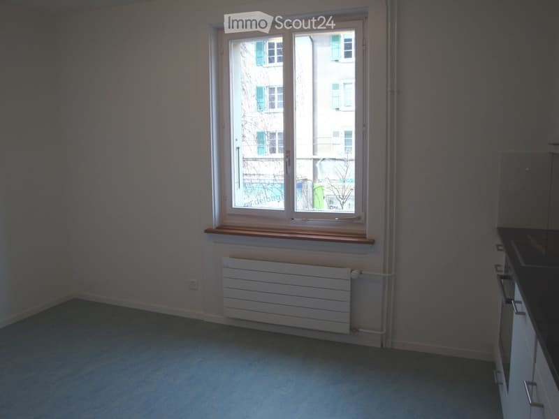 Empty room with light grey carpet, white walls, a window with wooden sill, radiator, and a small section of a kitchen counter visible on the right.