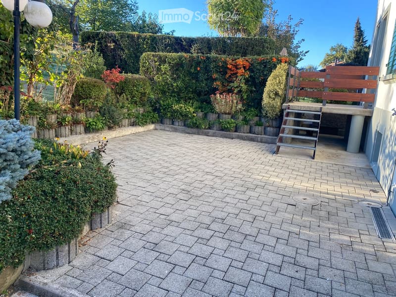 Well-maintained garden with a paved brick pathway, neatly trimmed hedges, flowering plants, and a wooden bench near the house.