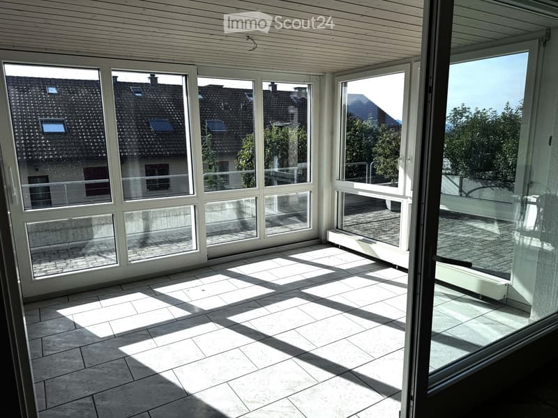 Bright, empty room with large glass windows and sliding doors leading to a terrace, tiled floor, and wooden ceiling beams.
