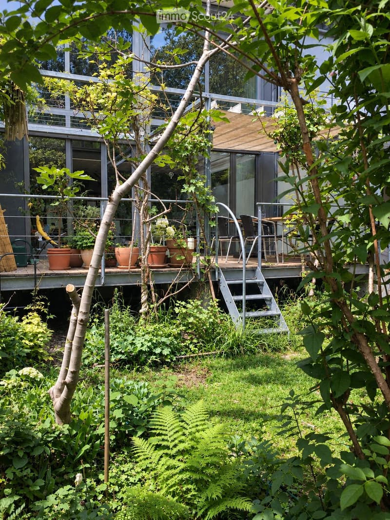 Garden area with lush greenery, a raised wooden deck, outdoor furniture, and potted plants. The deck is connected to a modern house with large glass windows and a staircase leading up to it.