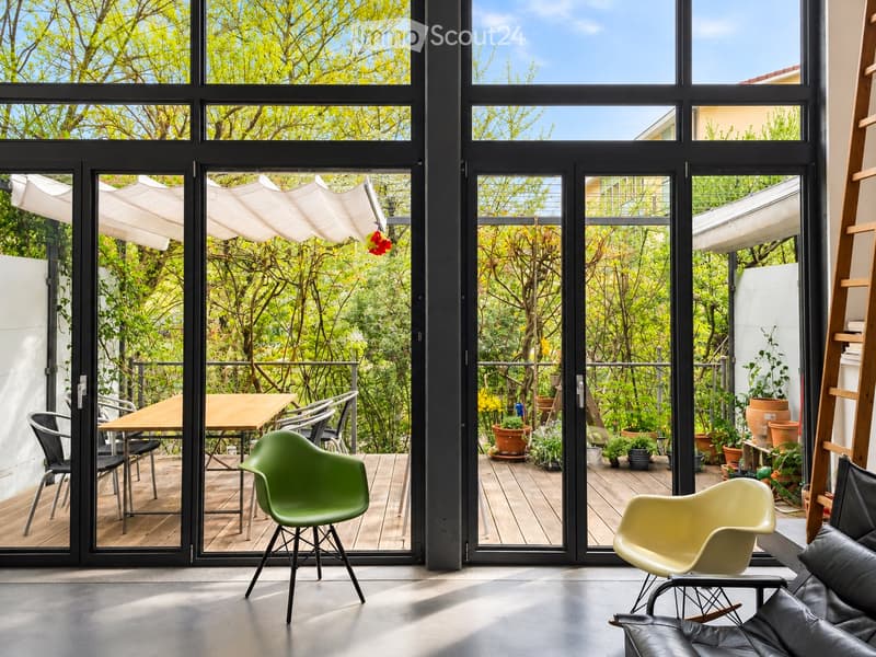 Modern living room with large glass doors opening to a balcony, featuring a green Eames chair, a beige Eames chair, a black leather sofa, and a wooden table with chairs on the balcony.