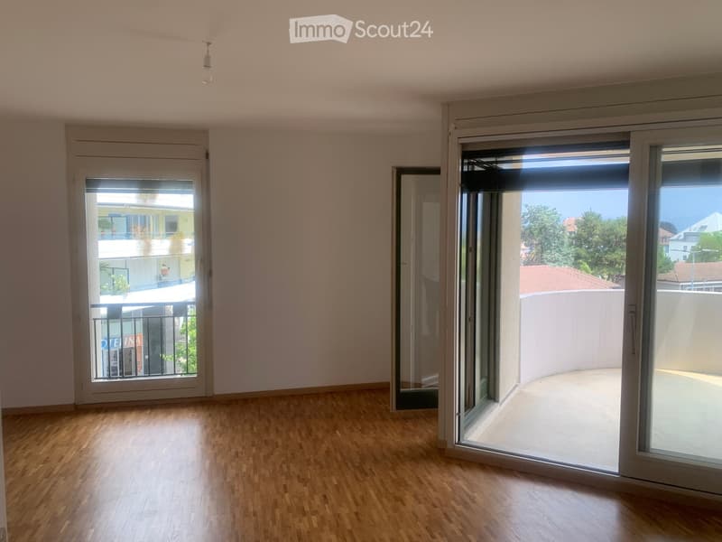 Empty room with wooden flooring, large sliding glass doors leading to a balcony, and a view of the surrounding area.