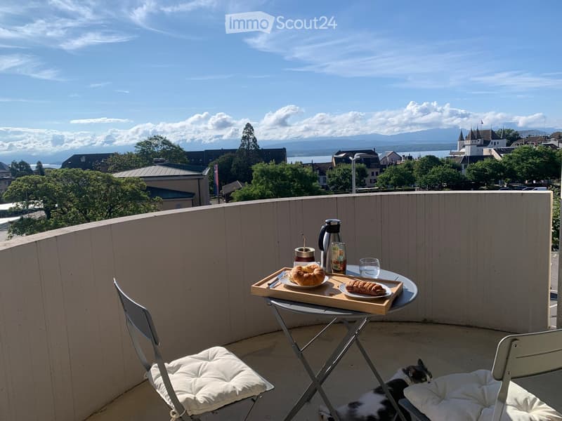 Balcony with a view of a town and lake, featuring a small table with breakfast items and two chairs with cushions, a cat resting nearby.