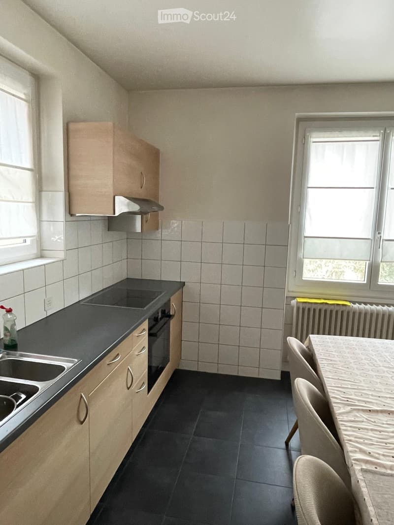 Modern kitchen with light wooden cabinets, black countertop, sink, oven, and a small dining area with a table and chairs.