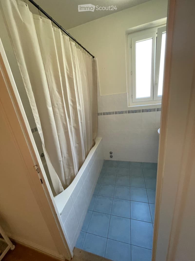 Bathroom with a bathtub, blue tiled floor, and a shower curtain partially drawn. A window is visible above the bathtub.