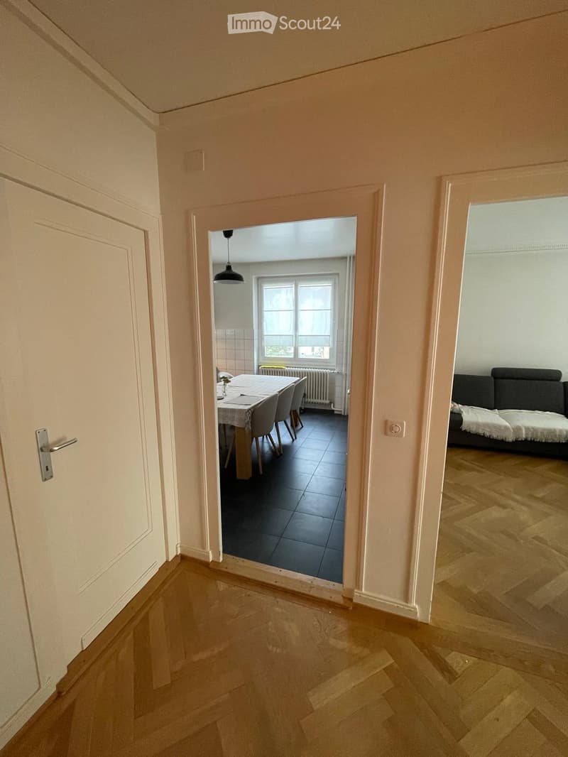 A hallway with light wooden flooring and white walls, leading to a dining area with a table and chairs, and a living area with a sofa and bed.