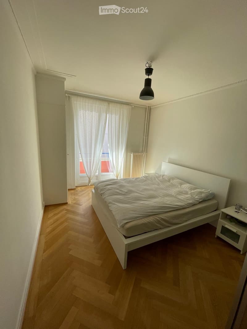 Minimalist bedroom with a white bed, nightstand, and wooden flooring. Large window with white curtains allows natural light to enter.