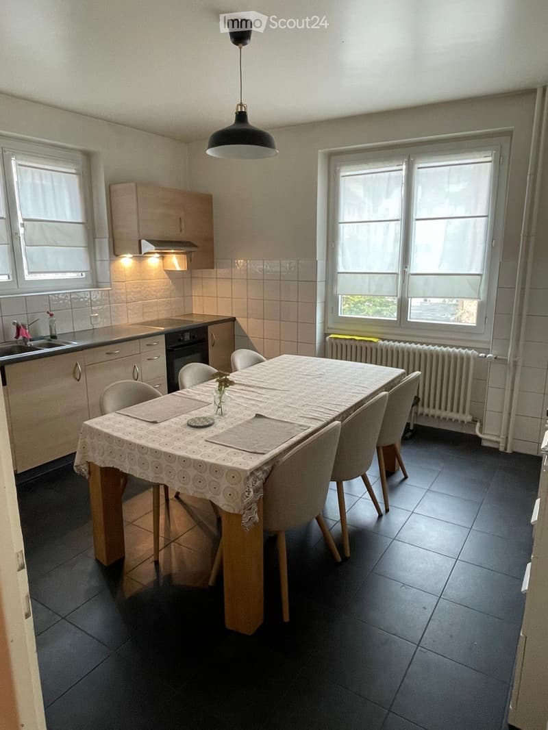Dining area with a table and chairs, cabinets, sink, and oven. The room has tiled walls and a dark tiled floor.