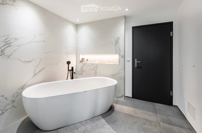 Modern bathroom with a freestanding bathtub, marble walls, a built-in shelf, and a black door.