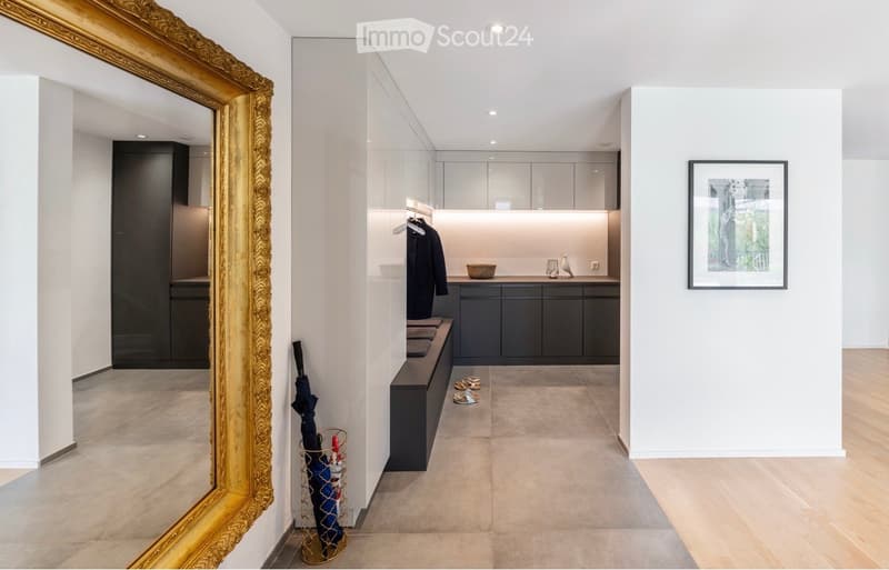 Modern hallway with a bench, coat rack, umbrella stand, and a small table with decorative items. The space features sleek, contemporary design with neutral tones and a large mirror on the left side.
