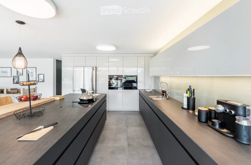Modern kitchen with white cabinets, stainless steel appliances, and a central island. Features include a coffee maker, knife block, pendant lighting, and a fruit basket on the counter.