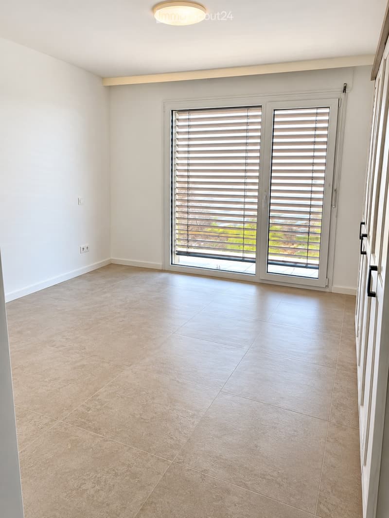 Empty room with beige tiled flooring, white walls, and large sliding glass doors with blinds leading to an outdoor area.