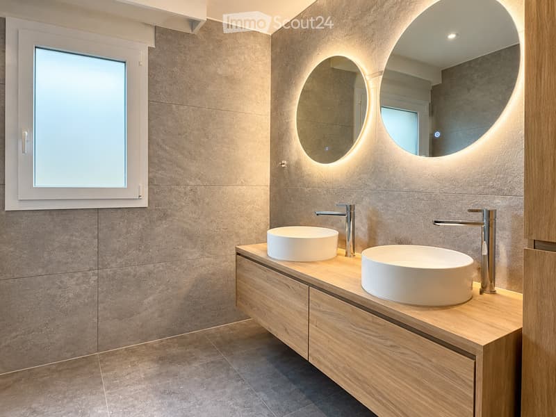 Modern bathroom with two white sinks on a wooden vanity, sleek chrome faucets, and a large illuminated round mirror above the sinks.