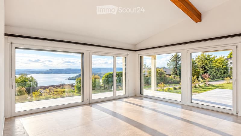 Bright, unfurnished room with large sliding glass doors offering panoramic views of a lake and surrounding landscape. The space features a high, slanted ceiling and light-colored tile flooring.