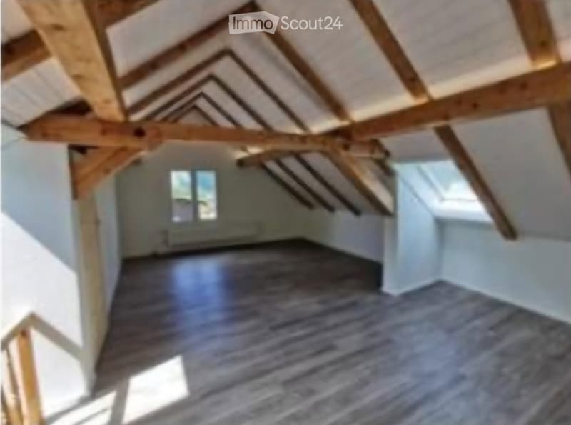Empty attic space with exposed wooden beams, wooden floor, and small windows allowing natural light.