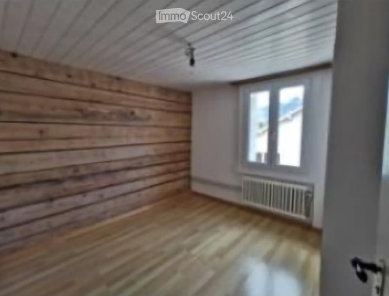 Empty room with wooden walls and floor, large window, radiator, and exposed wooden ceiling beams.