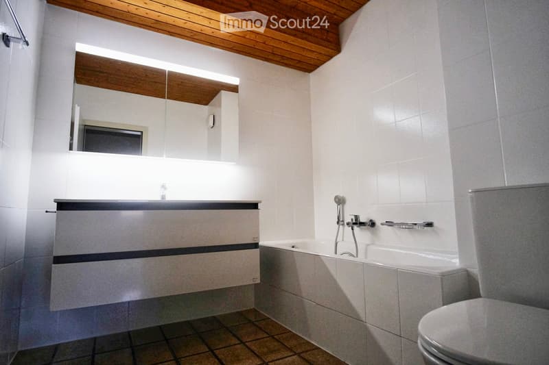 Modern bathroom with a wooden ceiling, white tiled walls, a large mirror above a vanity sink, a bathtub with a handheld showerhead, and a toilet.