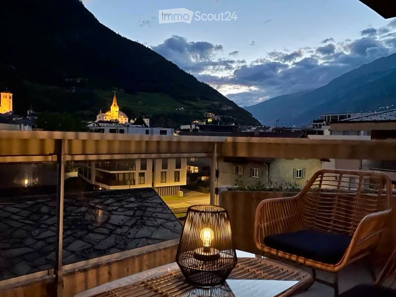 Balcony with a view of a town and mountains, featuring a wicker chair, small table, and a lit lamp.