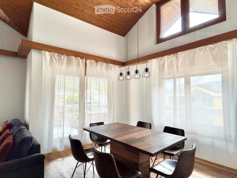 A modern dining area with a wooden table, several chairs, and a sofa, featuring large windows with white curtains and a wooden ceiling.