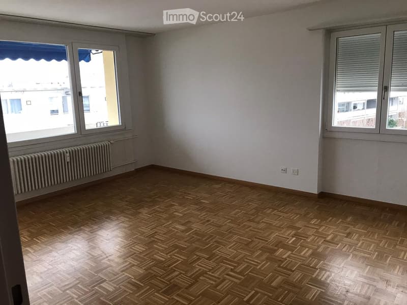Empty room with wooden parquet flooring, white walls, and large windows with blinds. Radiator visible under one window.