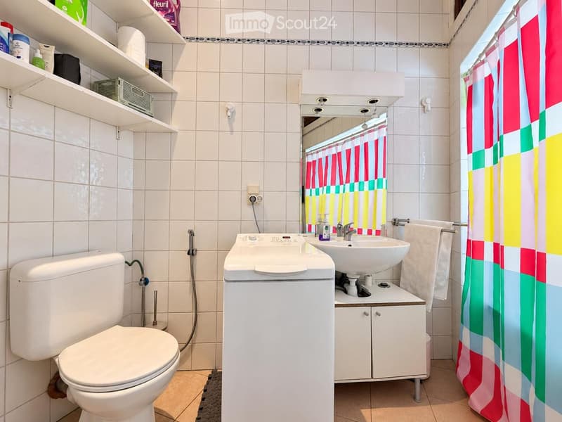 A compact bathroom with white tiles, a toilet, a sink with a mirror, a washing machine, and a colorful shower curtain.