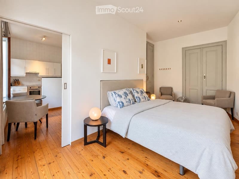 Cozy studio apartment with a double bed, nightstand, two armchairs, small table, and an open kitchen area with cabinets, refrigerator, and oven.